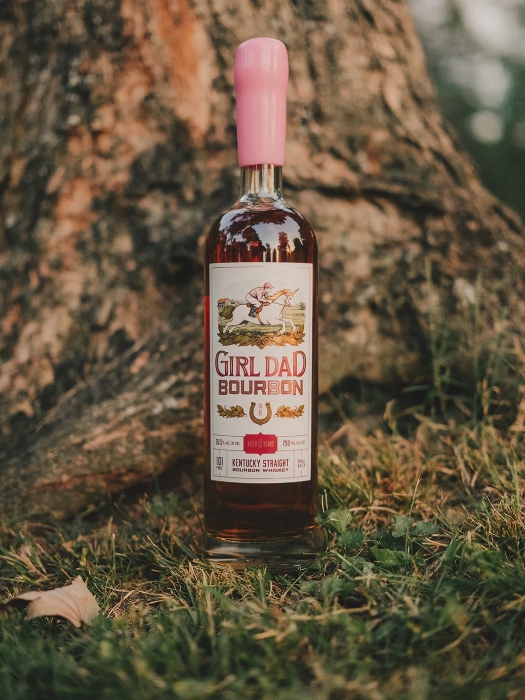 Girl Dad Bourbon 8-year bottle resting in the grass at the base of a tree